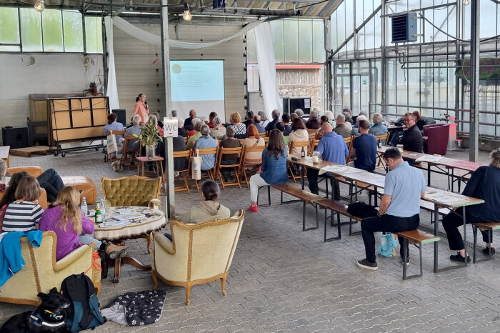 In einem alten Gewächshaus sitzen Menschen auf Stühlen und schauen auf eine Präsentation. Eine Frau steht vor der Leinwand uns spricht zu den Menschen.