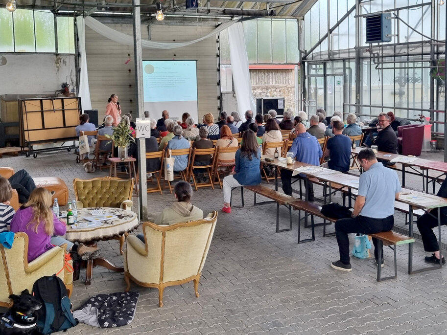 In einem alten Gewächshaus sitzen Menschen auf Stühlen und schauen auf eine Präsentation. Eine Frau steht vor der Leinwand uns spricht zu den Menschen.