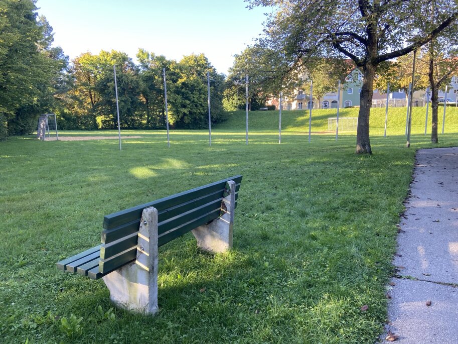 Auf einer Wiese steht eine Bank. Im Hintergrund ist ein Bolz-Platz zu sehen.