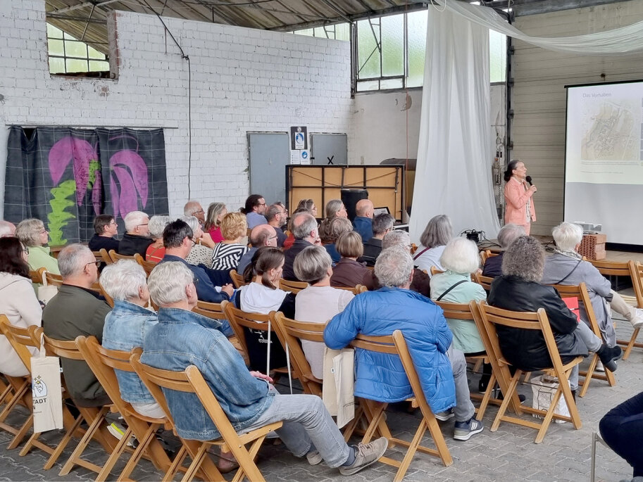Menschen sitzen auf Stühlen und schauen auf eine Präsentationsleinwand. Eine Frau erklärt die Grafik der Präsentation.